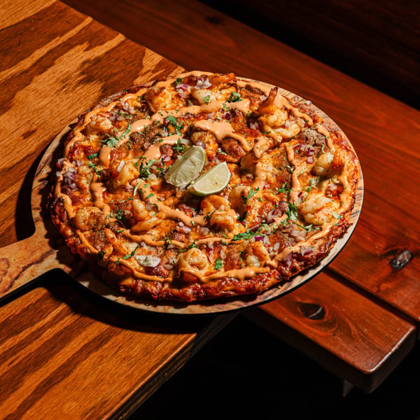 Indian shrimp pizza with sauce, red onion, cilantro, and lime wedges on a wooden paddle.