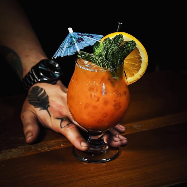Bartender's tattooed hand holding an orange cocktail garnished with mint, orange slice, and a blue umbrella.