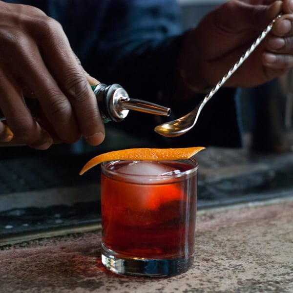 Bartender pouring a drink from a bottle into a spoon over a cocktail with an orange peel.