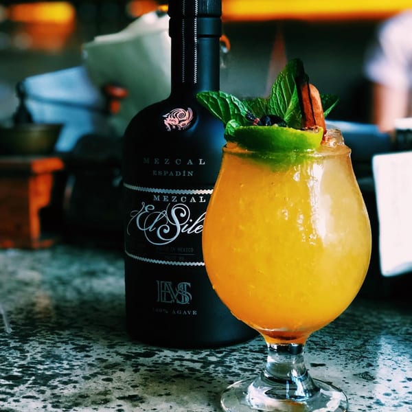 A vibrant orange cocktail garnished with mint and lime, next to a dark bottle of Mezcal.