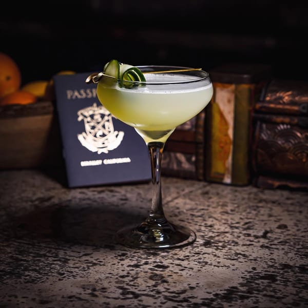 Light green cocktail with cucumber garnish, a passport, and oranges on a dark, textured surface.