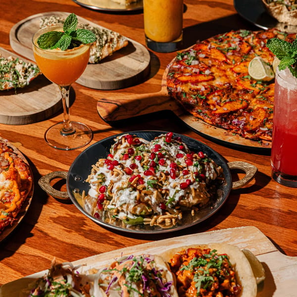 A wooden table laden with various Indian dishes, including pizzas, chaat, tacos, and cocktails.