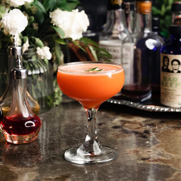 Orange cocktail with a green garnish on a bar, with bottles and white flowers in the background.
