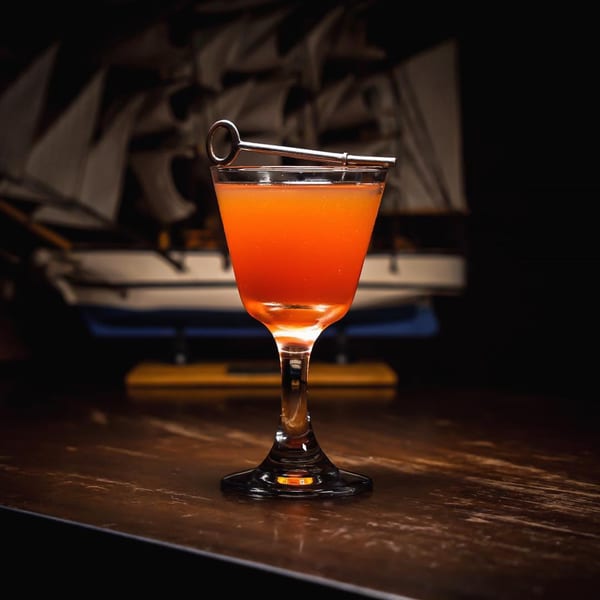 A vibrant orange cocktail in a coupe glass with a key garnish, on a wooden table.