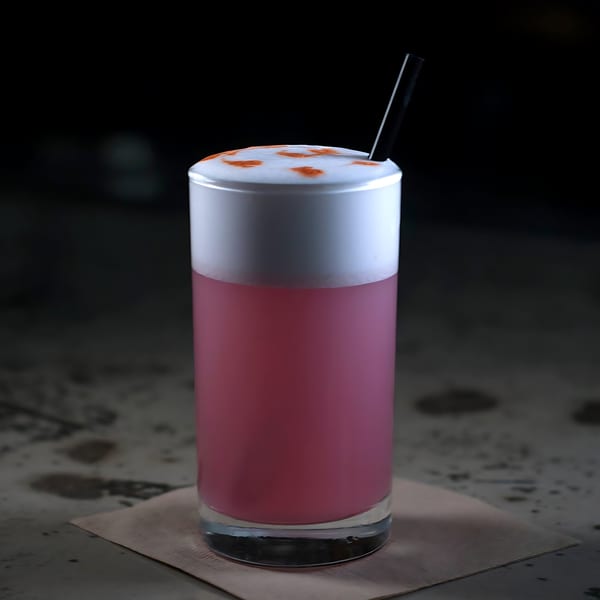 Pink cocktail with a thick white foam top, garnished with orange dots and a black straw.