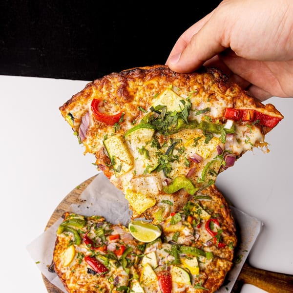 Hand lifting a slice of Indian pizza with bell peppers, zucchini, and cilantro from a wooden board.